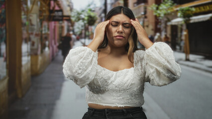 Fototapeta premium Woman with hand on forehead for headache on street wearing white embroidered blouse, wincing and rubbing temples; stress fatigue frustration.