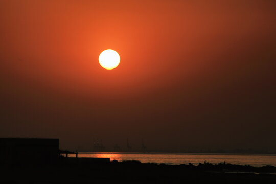 View of the radiant sun casting a warm glow over the tranquil sea, with dark silhouettes of buildings and rocks defining the horizon, Karachi, Sindh, Pakistan.