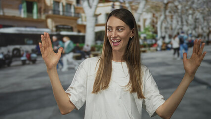 Fototapeta premium Woman with hands raised on street, palms open and smiling in a white tshirt with blurred bus and crowd behind her; joyful surprise.