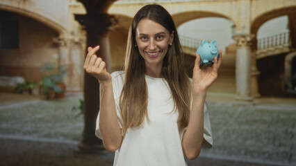 Young smiling woman holding blue piggybank and making finger heart gesture in building; savings...