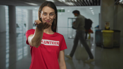 Woman volunteer extends hand palm up in airport terminal wearing red tshirt offering assistance;...