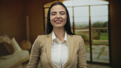 Fototapeta premium Young hispanic woman laughing and pointing indoors in a cozy living room setting with large windows in an apartment.
