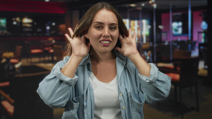 Young caucasian woman pressing hands to ears in restaurant, wincing at loud music and background...