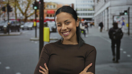 Woman smiling on busy street with arms crossed, exuding confidence and happiness in a vibrant urban...