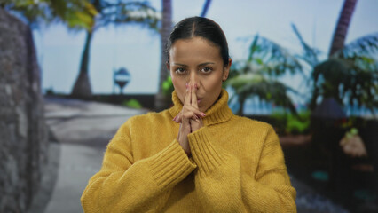 Woman wearing yellow sweater mimics shooting gesture with fingers outdoors amid palm trees on a sunny street.