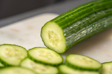 A whole cucumber rests on a cutting board with its freshly cut end in focus. Blurred slices in the foreground highlight the vegetable's juicy, crunchy texture.