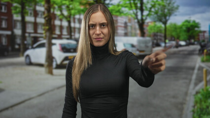 Young hispanic woman pointing index finger directly at camera on a city street with parked cars and stern expression; assertion boundary.