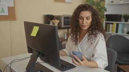 Woman holding smartphone while gazing at computer monitor at desk inside office building; focus efficiency.