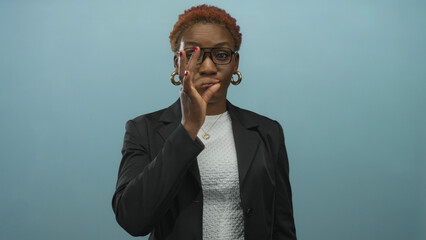 Woman in black blazer places finger to lips in bright blue studio background, emphasizing a sealed lip gesture with thoughtful expression  silence hush. © Krakenimages.com