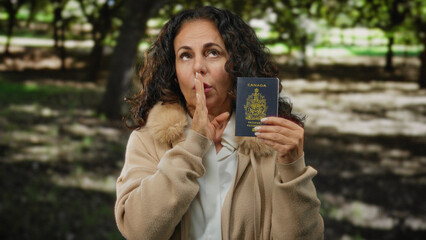 Middle-aged woman holding canadian passport while gesturing silence in a park setting, wearing a...