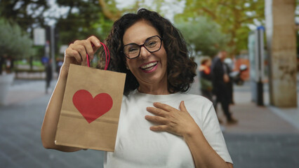 Woman outdoors holding a heart-decorated paper bag on a vibrant street, expressing surprise and...