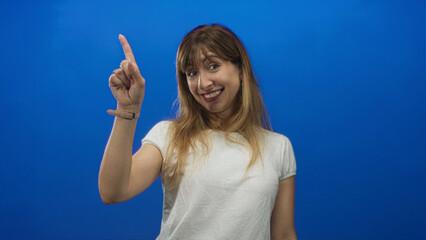 Young caucasian woman smiling and pointing index finger while wearing white t shirt in blue studio;...