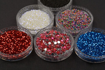 A group of six small, clear round containers holds various shiny nail art supplies, including red and blue glitter, white microbeads, colorful micro-caviar, and pink rhinestones