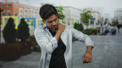 Young hispanic man rubbing neck and clutching shoulder while standing on a busy city street;...