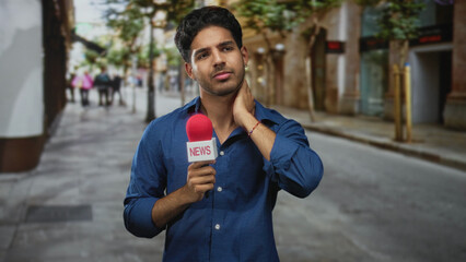 Man holding news microphone with red windscreen and touching neck on street, young hispanic reporter adjusting stance with closed eyes and subtle smile; pensive focus.