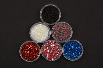 A top-down view of six clear containers filled with various colored glitters and beads, arranged in a triangular pattern on a black background