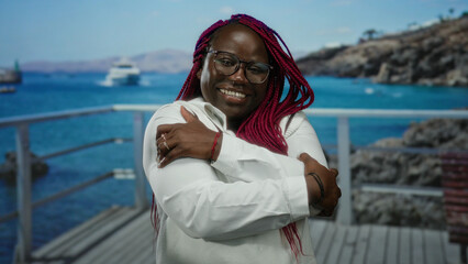 Woman with glasses and long braids smiling joyfully at the beachside, embracing herself against a backdrop of the sea and distant hills under a clear sky.