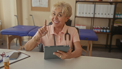 Senior physiotherapist with stethoscope tapping a tablet and pointing finger while seated at a desk in a medical building; supportive guidance.