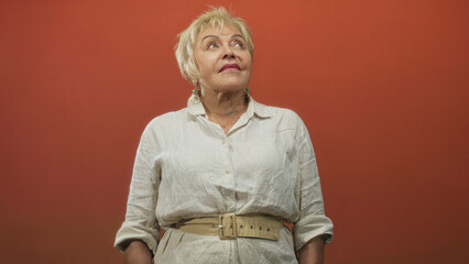 Senior woman in linen shirt with beige belt and dangling earrings gazes upward and to the side...