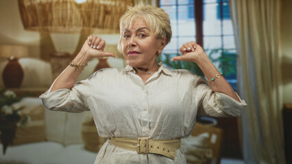 Senior hispanic woman with thumbs pointing to chest in studio bedroom set, wearing light blouse and...