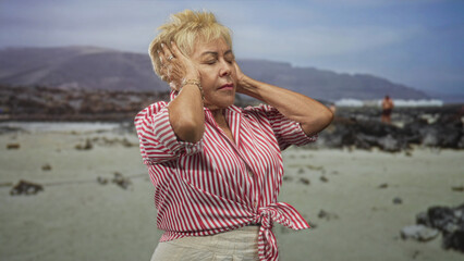 Senior woman covers ears with hands at a rocky beach, wearing red striped shirt tied at waist with...