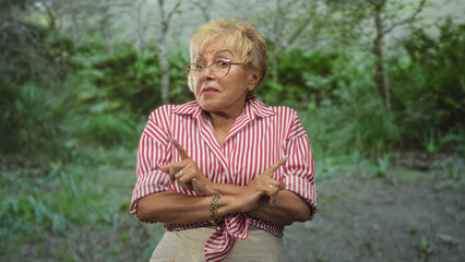 Woman pointing fingers with both hands in a forest, wearing a red striped tied shirt and glasses; playful defiance.