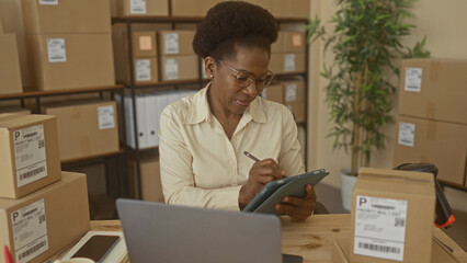 Woman handling cardboard boxes and tapping tablet with hand at a packing table in a building; focused small business efficiency.