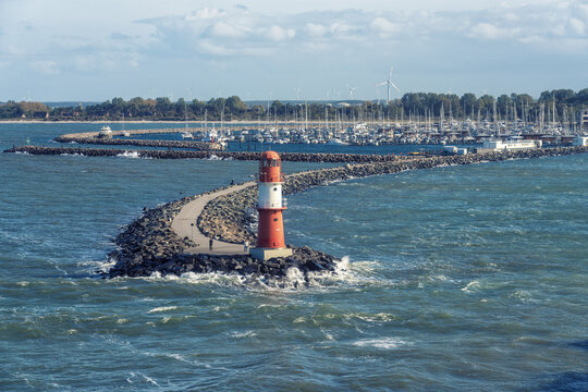 View of the gleaming red and white lighthouse standing proudly against the turbulent blue waters of the harbor, Rostock, Rostock, Germany.