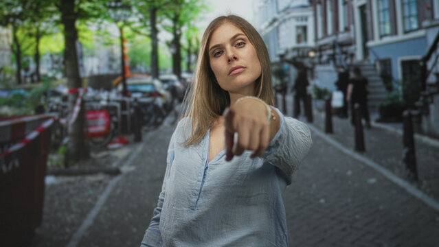 Young blonde woman stands amid parked bicycles on narrow cobblestone street flipping middle finger; defiance.