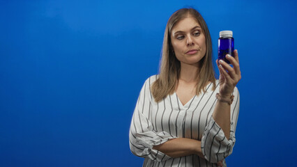 Woman with thoughtful expression pointing to prescription pill bottle against bright blue studio wall; doubt health.
