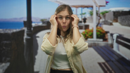 Fototapeta premium Woman with hands pressing temples in bright seaside street wearing casual attire and thoughtful posture; calm reflection introspection mood.