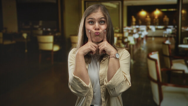 Blonde woman with fingers on cheeks pouting inside a warm lit restaurant dining area crowded with empty chairs; playfulness.