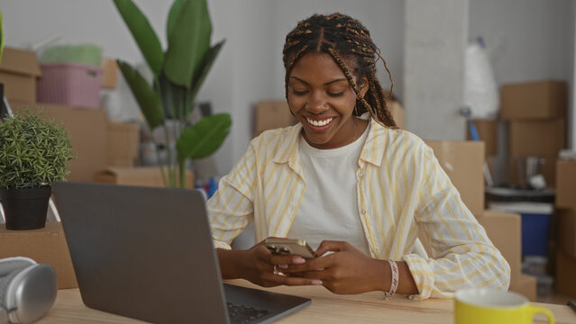 African american woman smiling and typing with hands on phone and laptop in building with moving boxes; small business startup happiness.