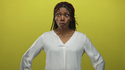 Woman with braided hair wearing a white blouse posing akimbo in yellow studio with hands on hips and a puckered expression; playful defiance.