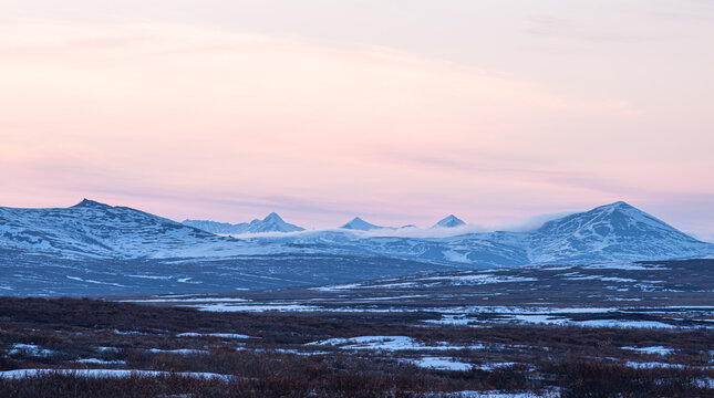 View of snow-dusted peaks stand silhouetted against a pastel sky, the frozen tundra stretching out in muted browns and whites, a tranquil vista, Nome, Alaska, United States.