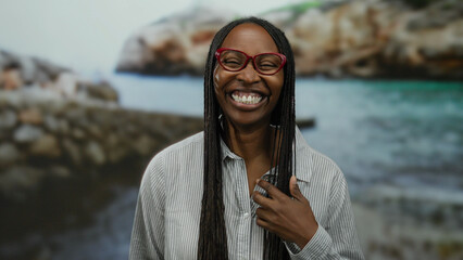 Smiling woman with glasses and braided hair at a seaside location, showcasing an inviting expression against a scenic beach backdrop.
