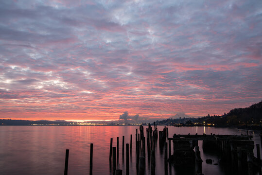 View of a serene sunrise with the pink and purple hues painted across the cloudy sky reflected on the calm water near wooden structures, Tacoma, Washington, United States.