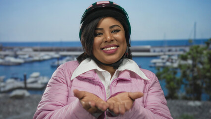 Woman standing at seaside port wearing helmet and pink jacket extending hands to camera with a backdrop of boats and the blue sea.