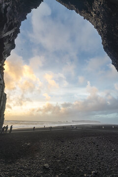 View of black sand beach framed by basalt columns under a sky streaked with sunset hues, casting long shadows on the shore, Reynisfjara Beach, Vik, Iceland.