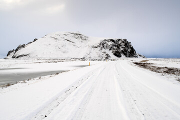 View of a snow-laden road stretching towards a dark, rugged mountain under a pale sky, a stark contrast of light and shadow in the winter landscape, Reynisfjara Beach, Vik, Iceland.