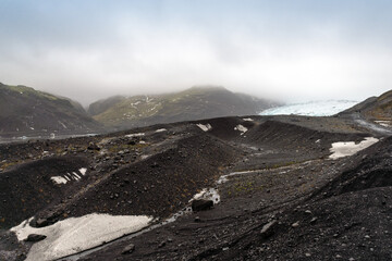 View of black volcanic sand contrasting with snow-touched earth, mountains shrouded in mist, and a pale sky in Reynisfjara Beach, Vik, Iceland.