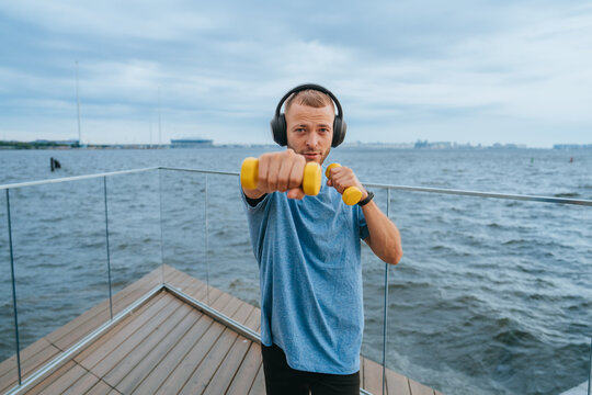 A man wearing headphones with a resistance band wrapped around his shoulders stands on a deck, focused on his phone amidst an oceanic tableau.