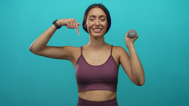 Young hispanic woman pointing to toned arm while holding a 1kg dumbbell in a teal studio; confidence wellness health.