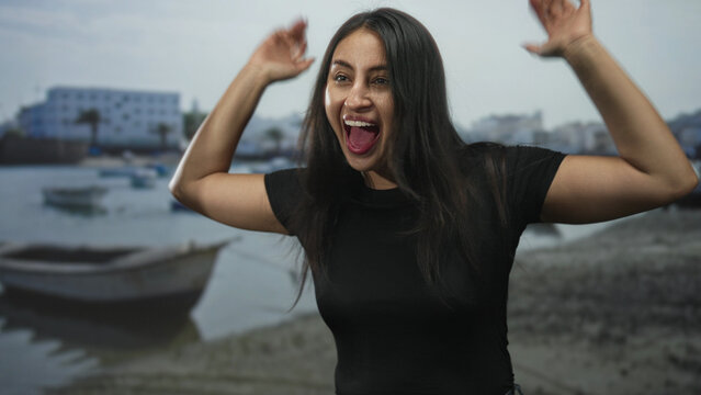 Woman raising arms and cheering on rocky beach with boats and harbor buildings visible; joy celebration. - Powered by Adobe