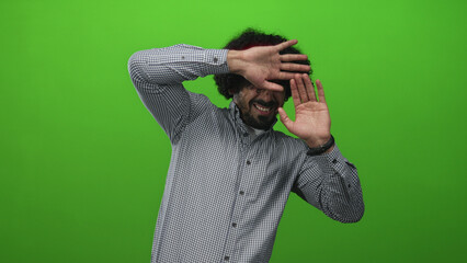 Man with curly hair and beard reaching forward with both palms toward camera in bright green studio wearing checkered shirt; fear tension alarm.