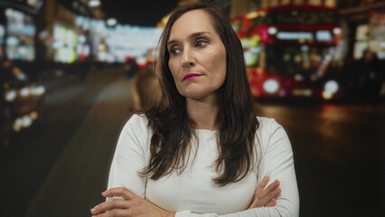 Woman standing on busy street at night with double-decker bus in background, arms crossed, serious...
