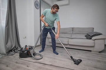 Man vacuuming floor and cleaning home interior. Person doing household chores with vacuum cleaner in living room. Concept of domestic work, home hygiene, and clean lifestyle