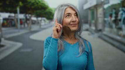 Senior woman with grey hair in a blue sweater stands thoughtfully on a bustling street, pointing upwards with curiosity and confidence in an outdoor urban setting.