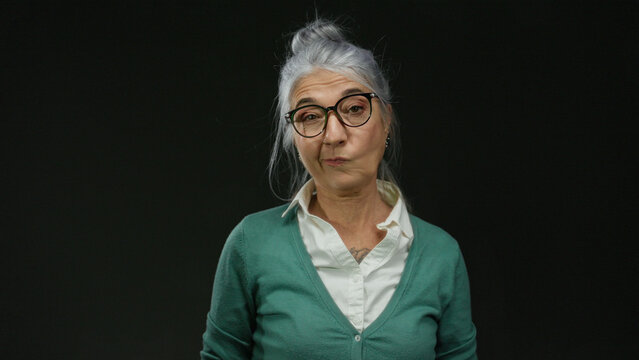 Senior grey-haired woman with glasses posing against a black background wall, expressing a contemplative mood with her arms crossed.