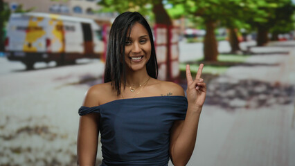 Woman smiling and making peace sign outdoors on a city street, exuding happiness and confidence on a sunny day with trees and a colorful van in the background.